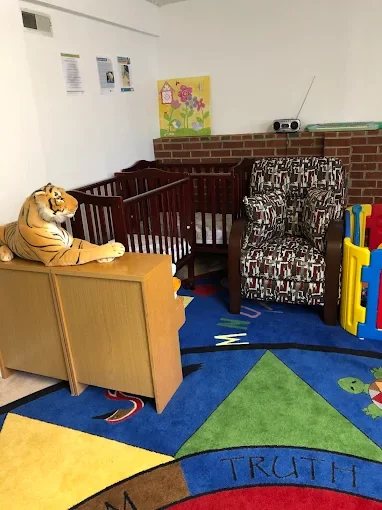 A stuffed tiger toy placed on a wooden cabinet near a crib in a colorful children's playroom with a patterned armchair, a play fence, and a vibrant alphabet-themed rug.