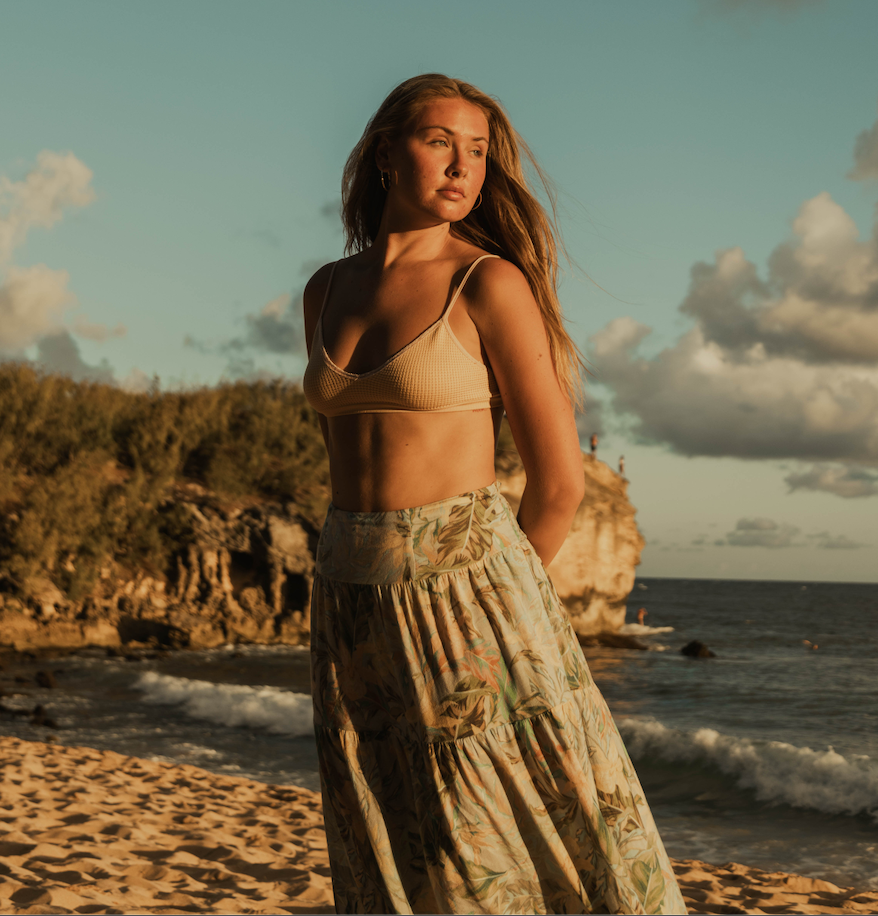 A DFW based florist stands on a sandy beach with rocks and cliffs in the background.