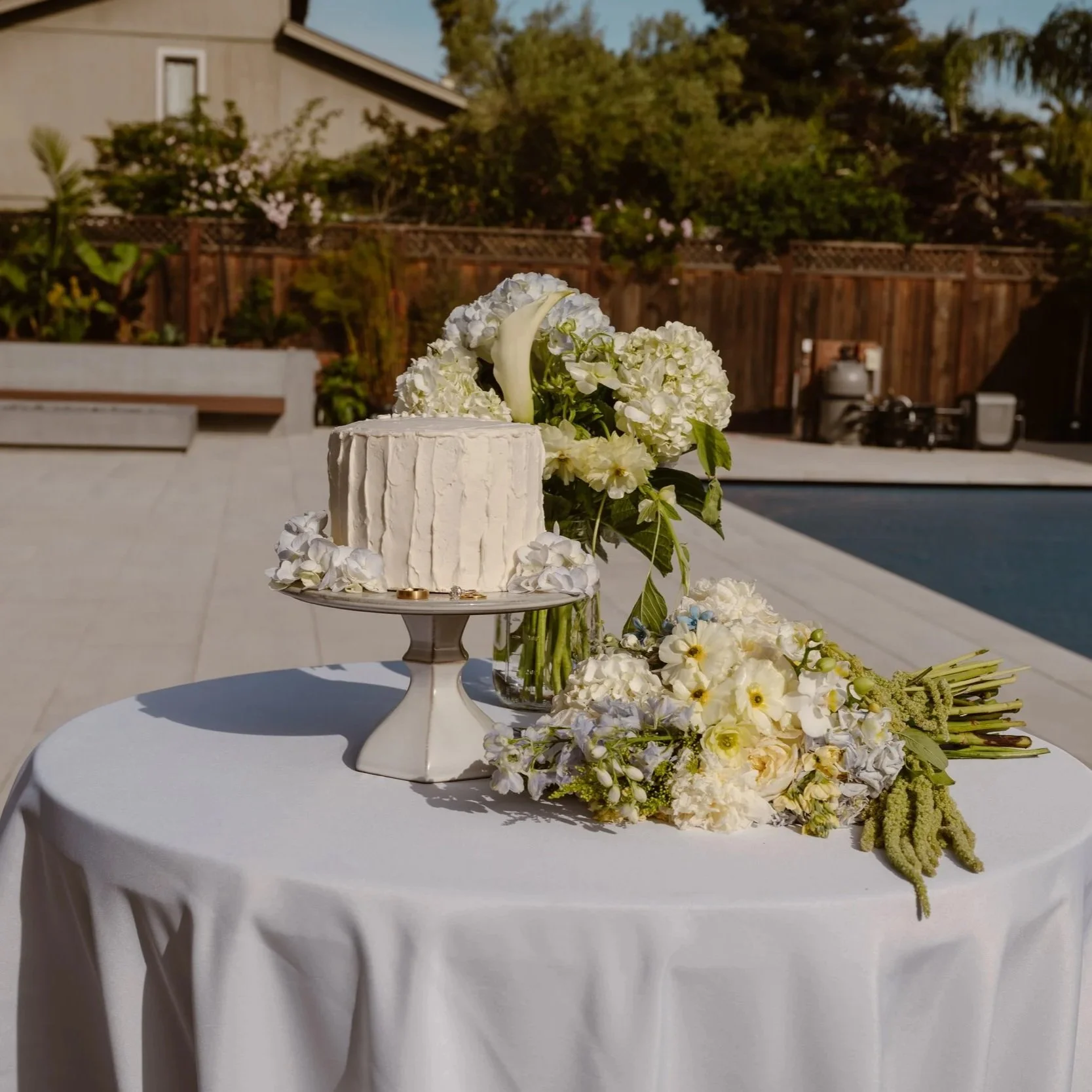 A white cake decorated with white flowers, including calla lilies and hydrangeas, placed on a round table covered with a white tablecloth, located in Prosper, Texas.