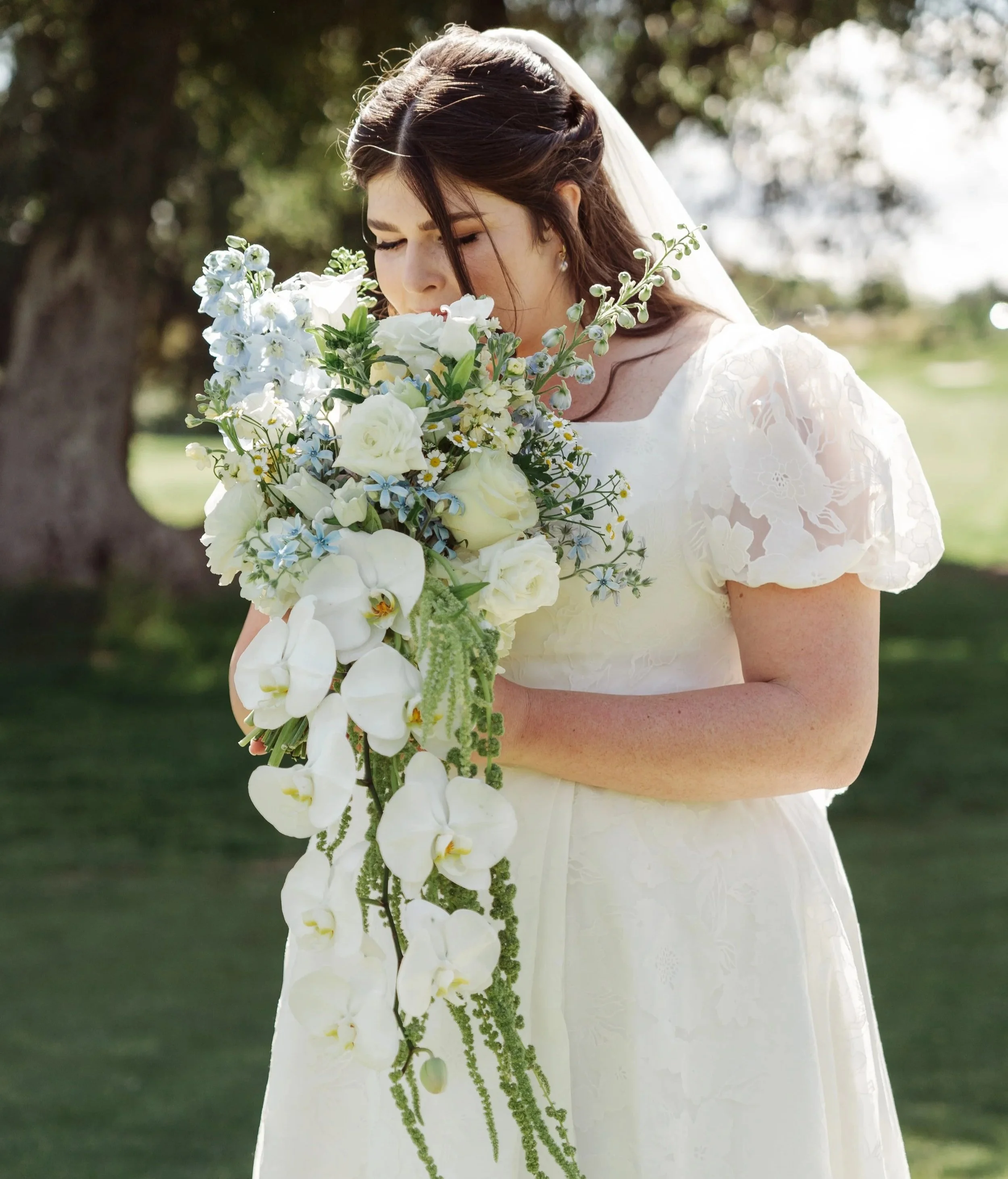 A woman in a white wedding dress holds a cascading bouquet of white and blue flowers in DFW, Texas.