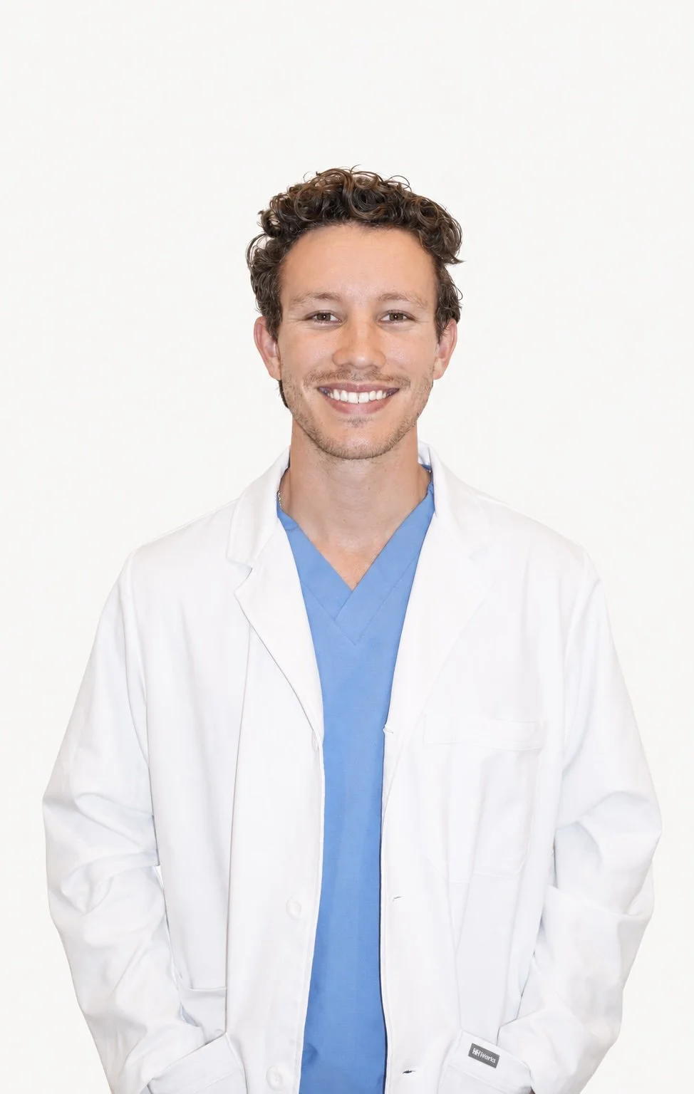 A smiling male doctor in a white coat and blue scrubs standing against a plain white background.