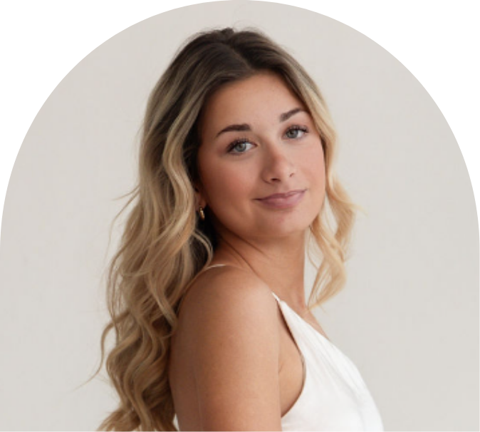 A young woman with long, wavy blonde hair and light skin looking over her shoulder with a gentle smile, wearing a sleeveless white top.