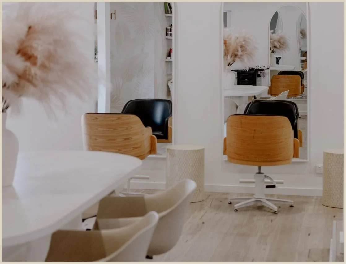 A modern, minimalist salon with mirrors, wooden and black salon chairs, and decorative elements such as a fluffy pampas grass arrangement and textured white baskets.