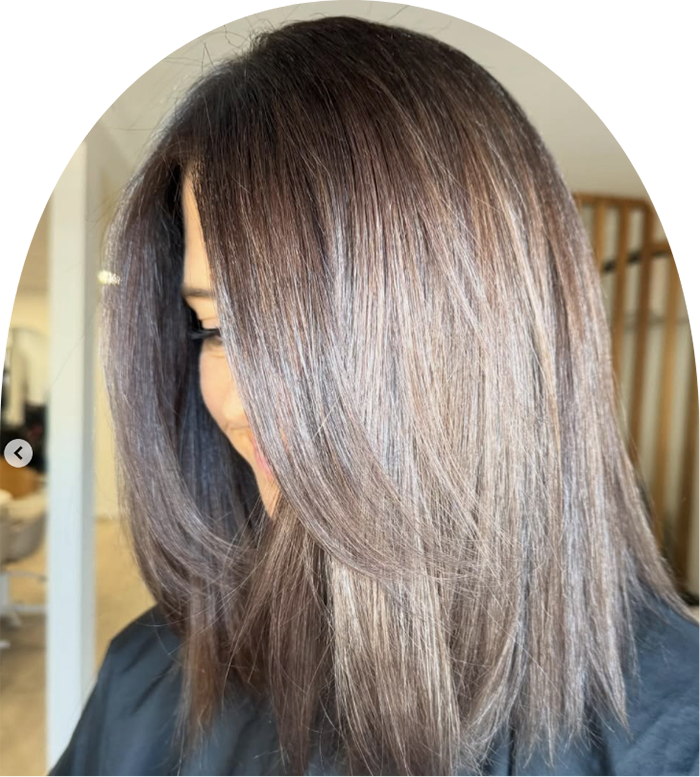 A woman with straight, shoulder-length, dark brown hair with lighter highlights, looking downward in a salon setting.
