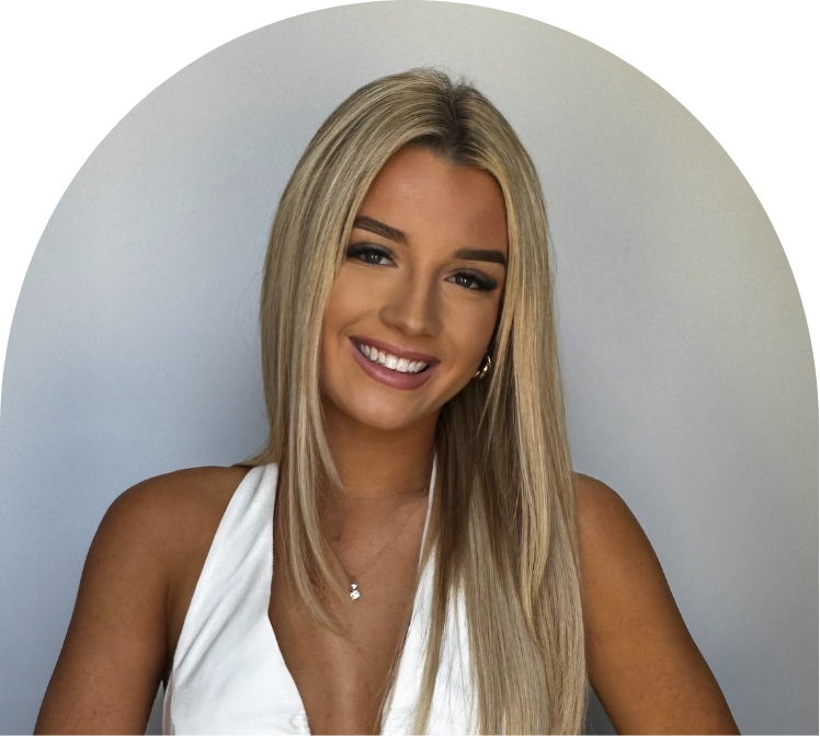 A young woman with long blonde hair, smiling, wearing a white sleeveless top and a necklace, standing against a light-colored wall.
