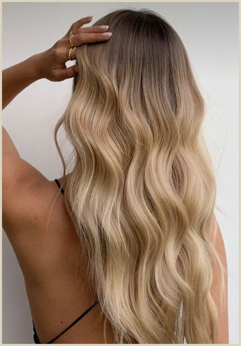 Back of a woman's head with long, wavy, blonde hair and hand touching her hair.