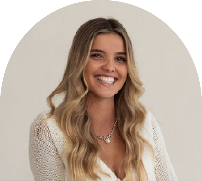 Smiling woman with long wavy blonde hair wearing a white top and layered necklaces, standing against a plain light background.