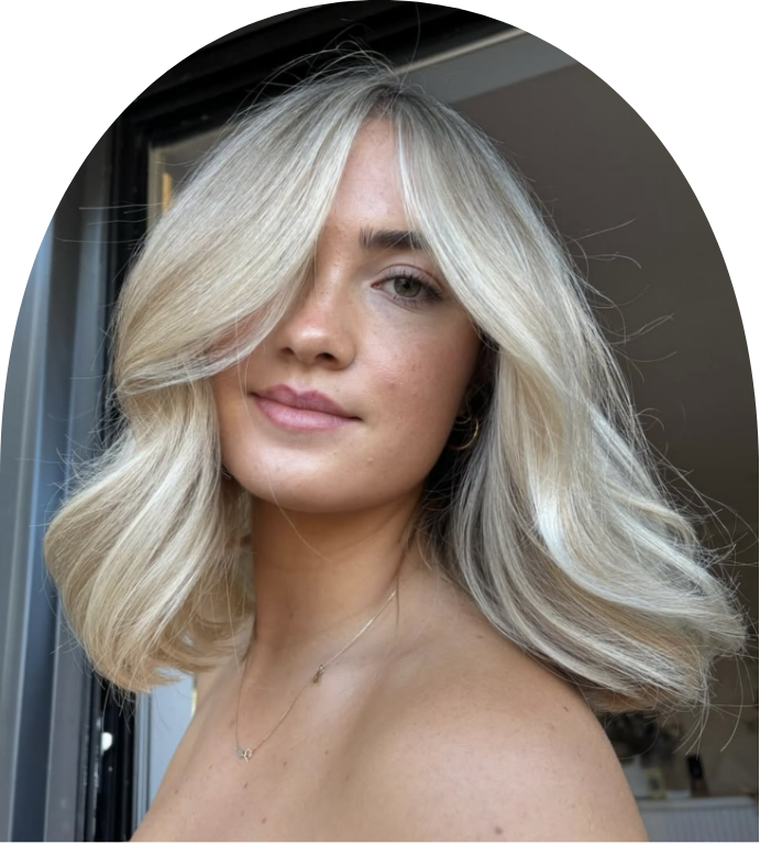 A young woman with wavy blonde hair and light skin taking a selfie inside a room, with a window and curtain visible in the background.