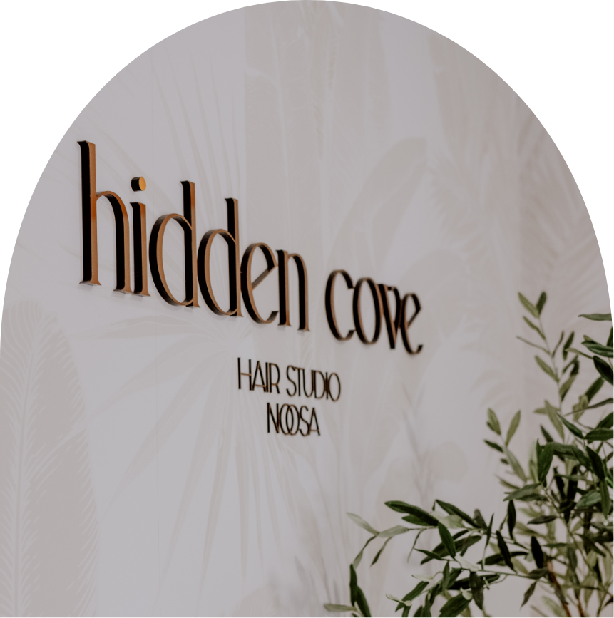 Sign on a wall with the text 'hidden core' in large, 3D letters and 'HAIR STUDIO NOOSA' underneath in smaller text, with a plant partially visible in the foreground.