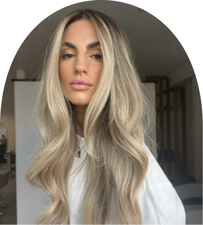 A young woman with long, wavy blonde hair, full lips, and well-defined eyebrows, wearing a white top, posing indoors.