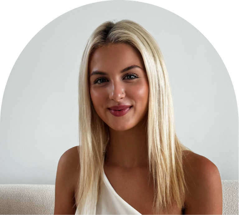 A young blonde woman smiling, wearing a white sleeveless top against a plain background.