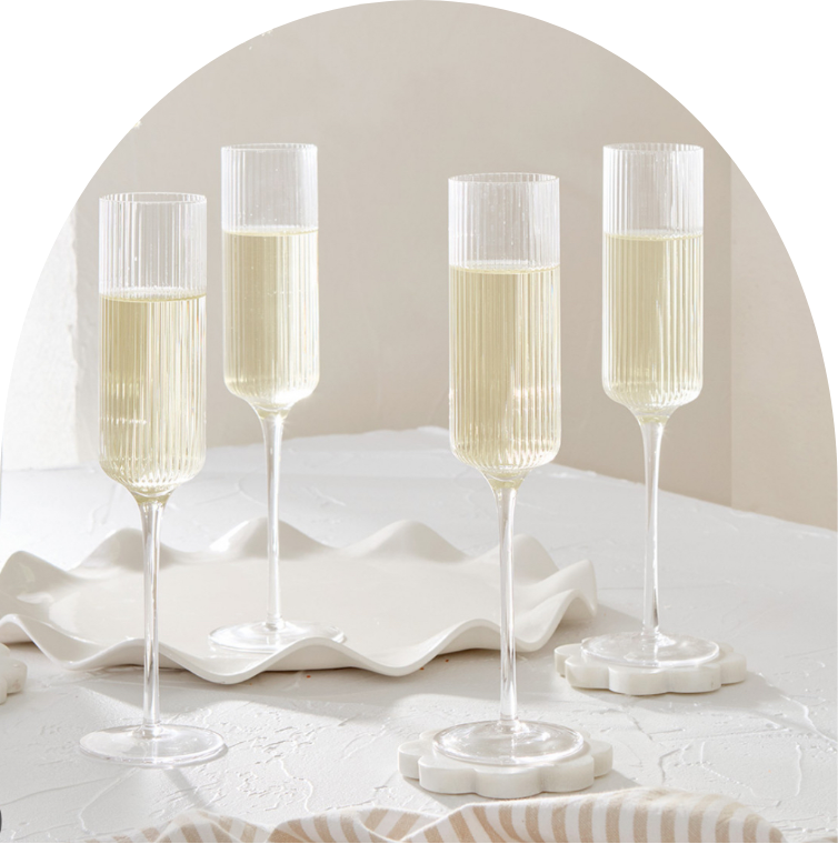 Four champagne glasses filled with a clear sparkling beverage, arranged on a white textured surface with a minimalist white dish and a striped cloth nearby.