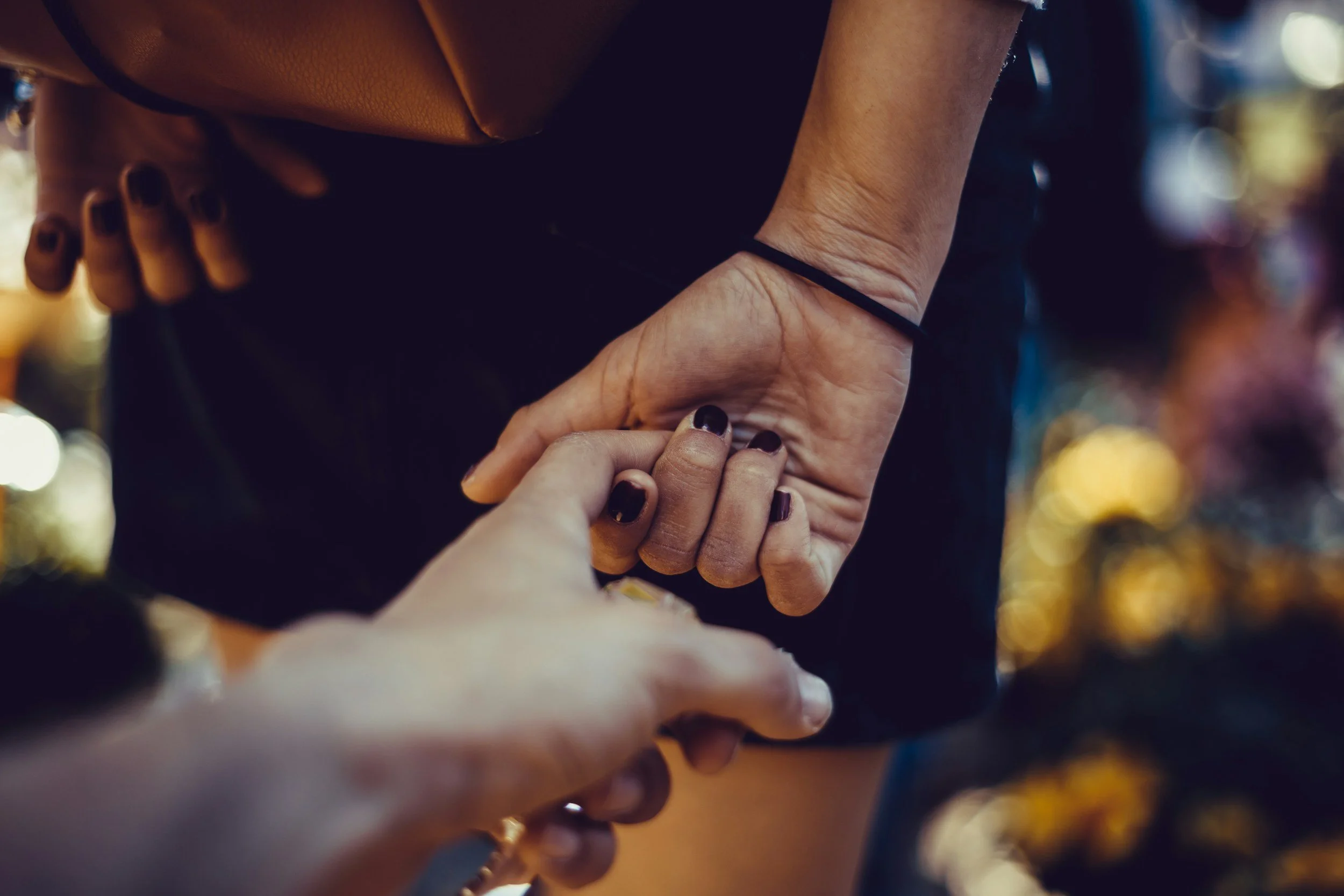 Close-up of two hands holding each other, with one person's hand grasping the other's wrist. The background is blurry with warm, colorful lights.