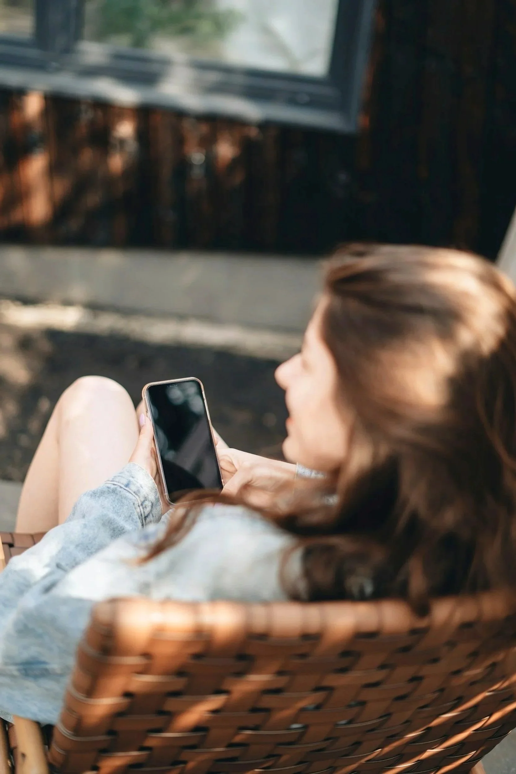 A woman with brown hair sitting on a wicker chair, looking at her smartphone outside near a wooden wall and window.