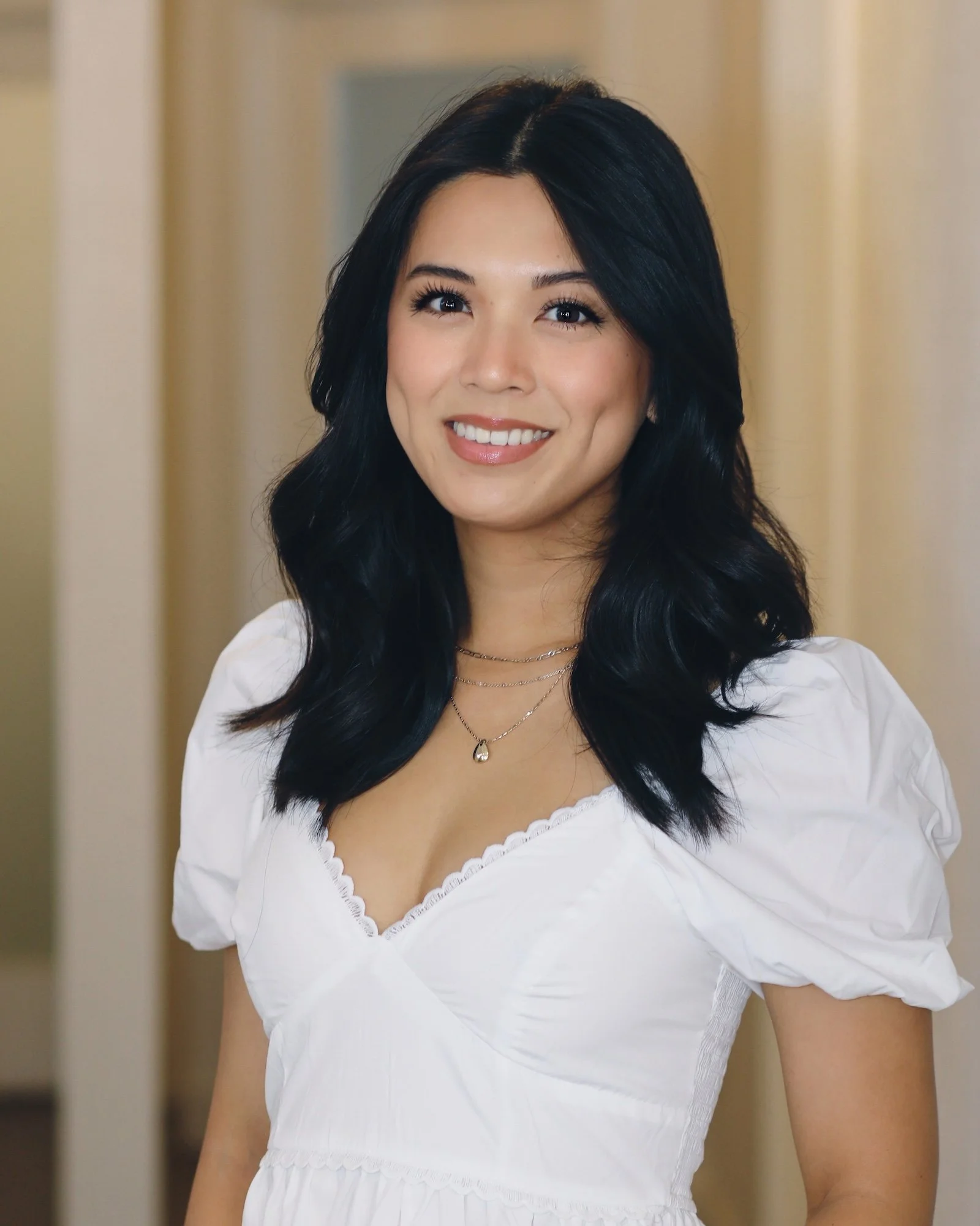 Young woman with long black hair wearing a white blouse and layered necklaces, smiling at the camera indoors.