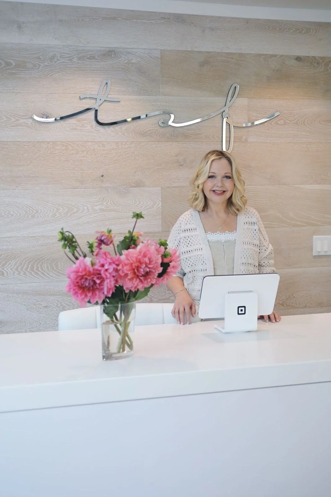 A woman with blonde hair behind a white reception desk with a vase of pink flowers, in front of a wood-paneled wall with a metallic sign that says "TSF."