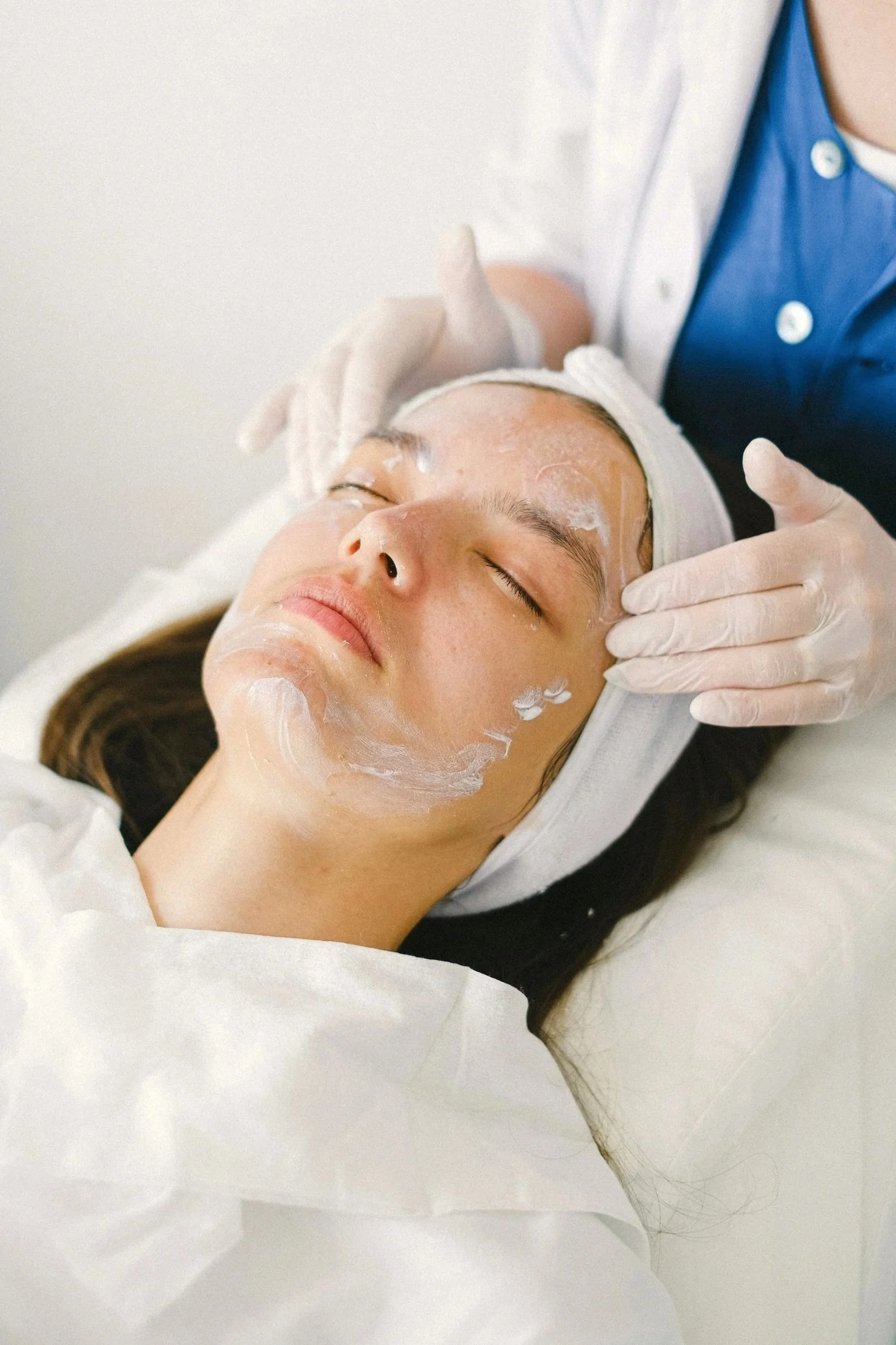 A woman receiving a facial treatment with a skincare product applied on her face, lying down with her eyes closed, in a spa or skincare clinic setting.