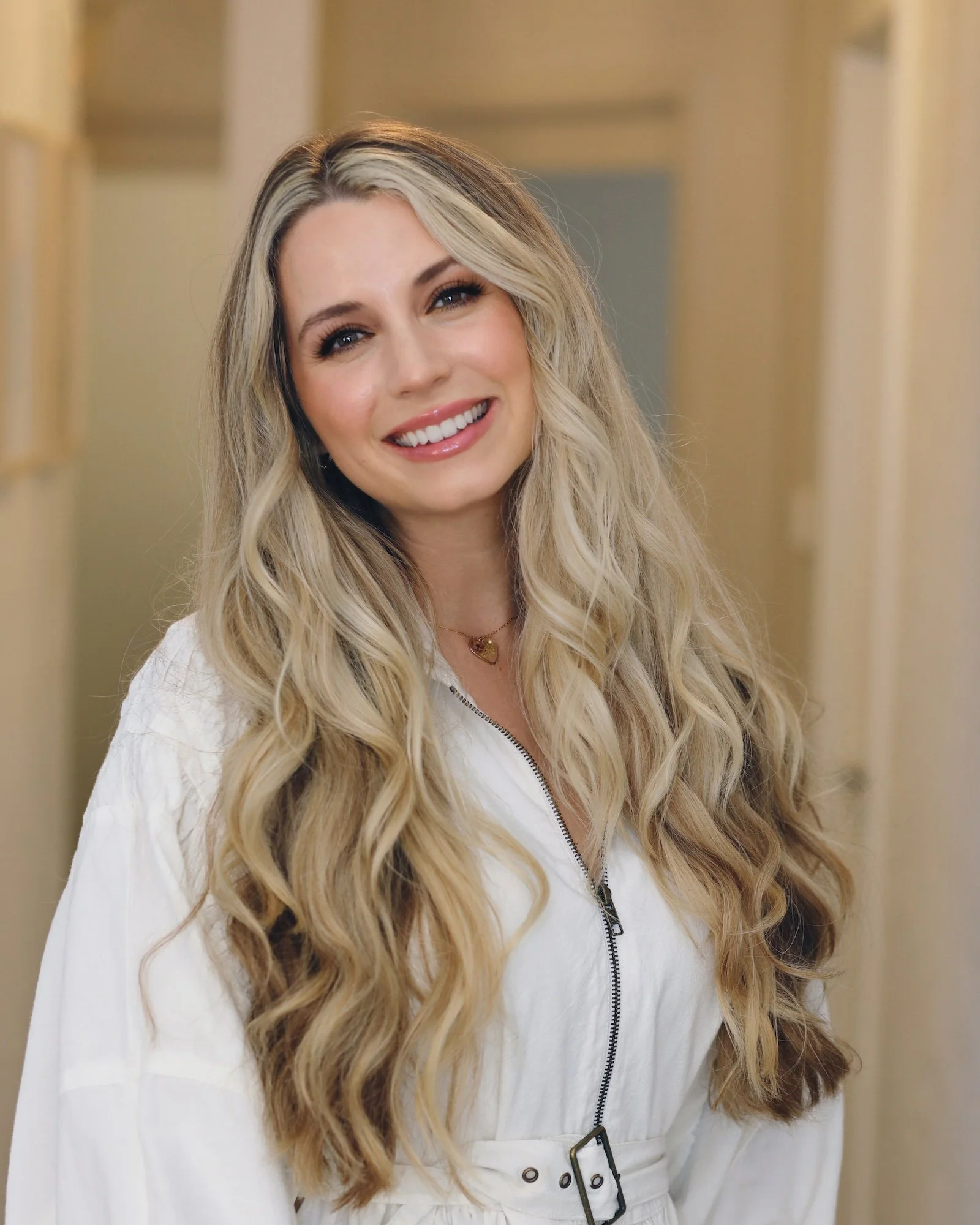 A woman with long, wavy blonde hair and light skin smiling while looking at the camera, wearing a white zip-up jacket and a heart-shaped necklace, standing in an indoor setting.
