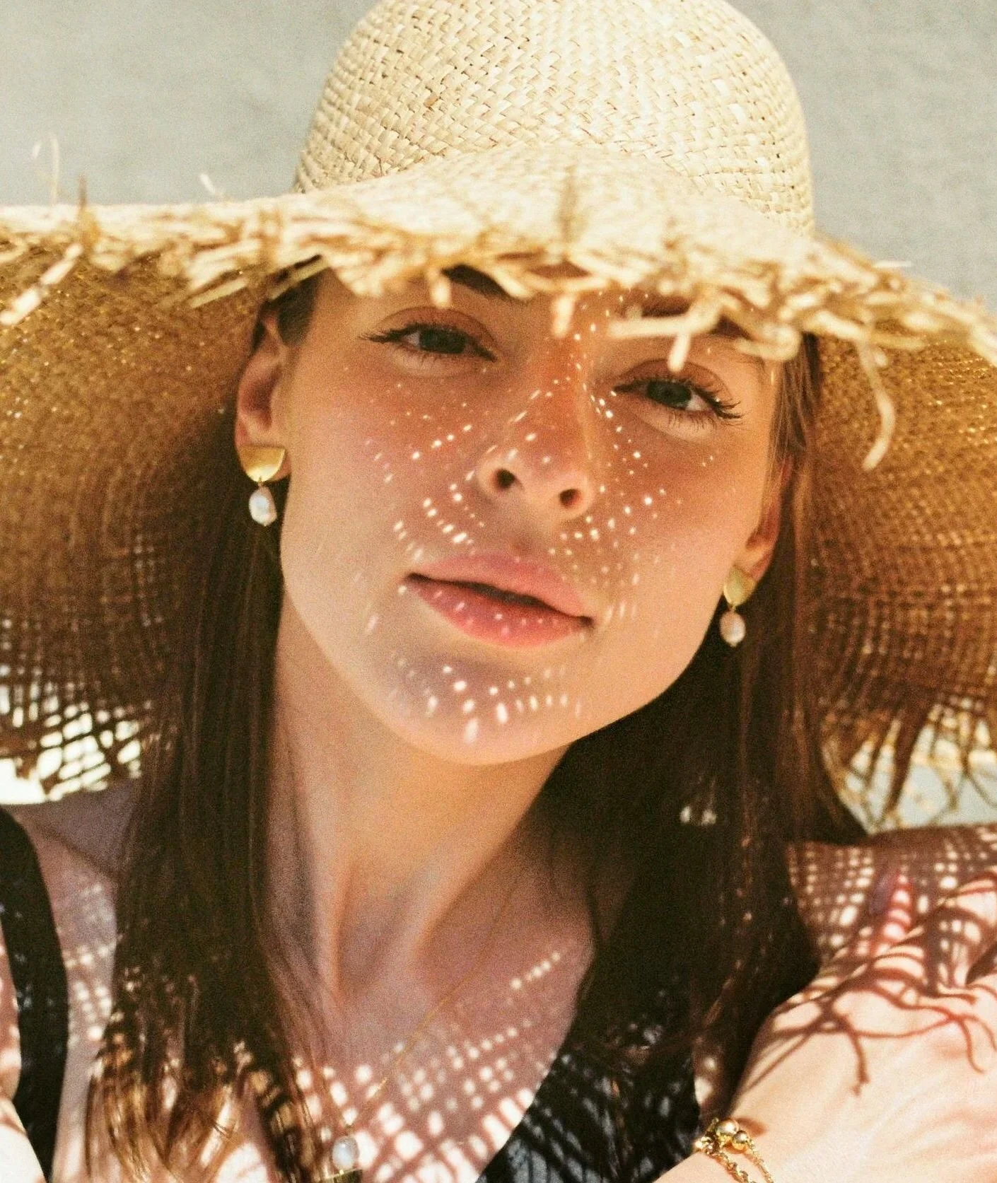 Woman wearing a wide-brimmed straw hat and earrings, with sunlight creating dotted shadows on her face.