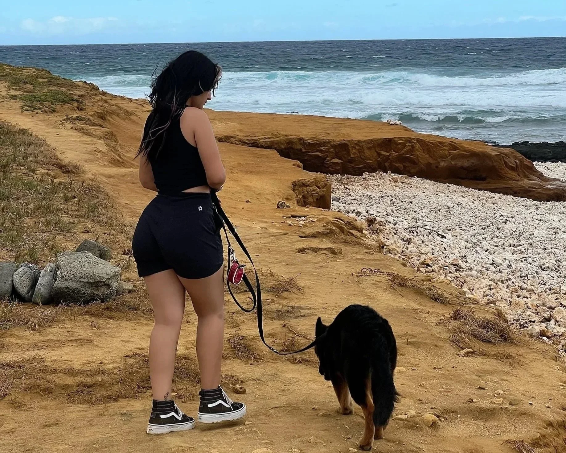 A woman standing on a sandy trail near the beach with a black dog on a leash, overlooking the ocean with waves crashing onto rocks and a cloudy sky.