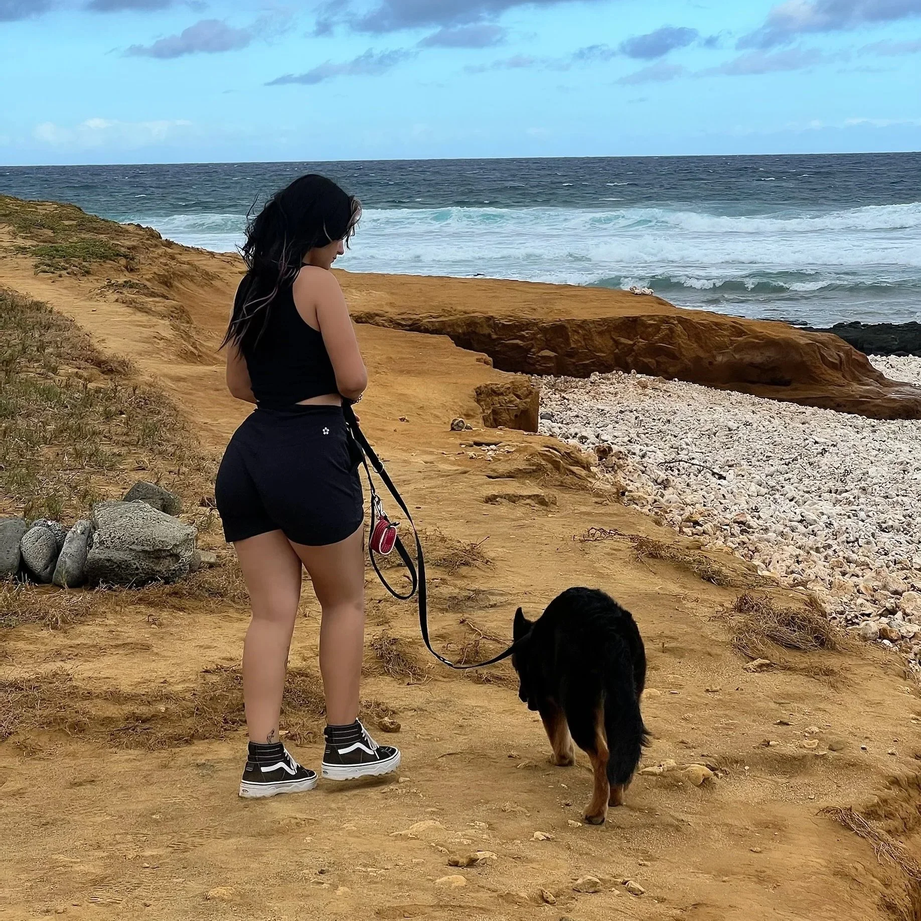A woman with black hair, dressed in a black top, black shorts, and black sneakers, holds a leash attached to a black and brown dog on a sandy cliffside overlooking the ocean with waves and a partly cloudy sky.