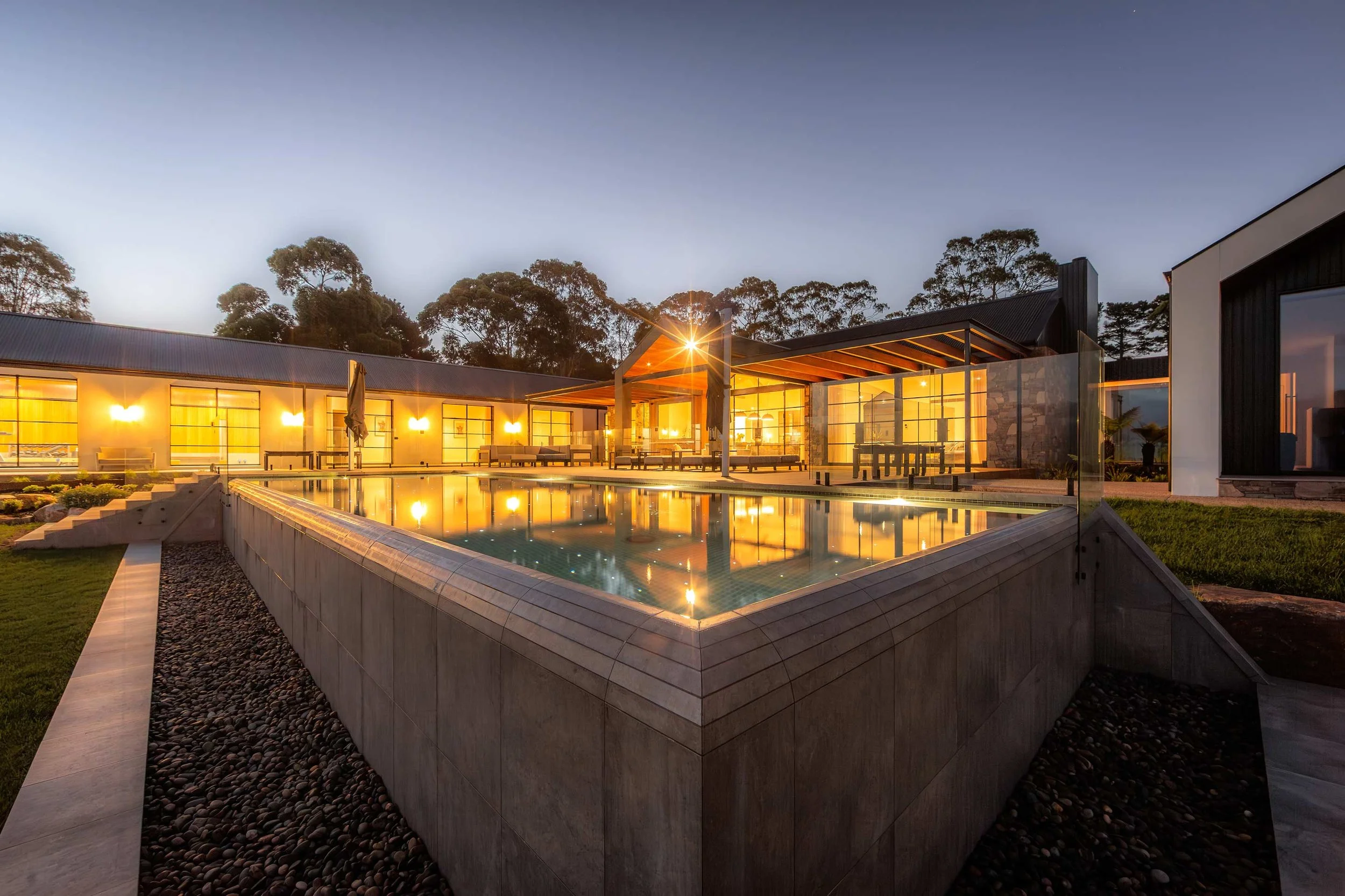 A wet-edge swimming pool illuminated with subtle, starlight-like lighting, surrounded by a warm and beautifully lit modern home.