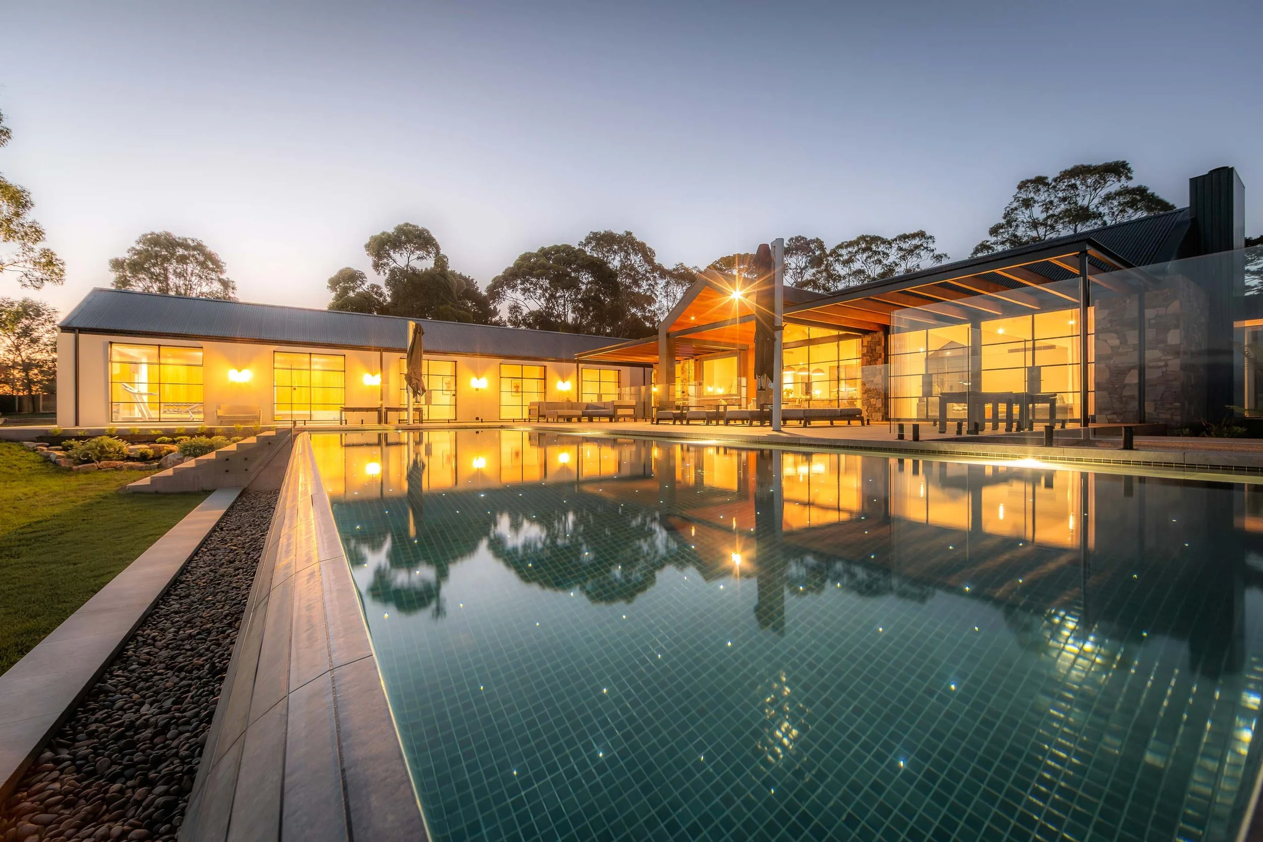 A wet-edge swimming pool illuminated with subtle, starlight-like lighting, surrounded by a warm and beautifully lit modern home.