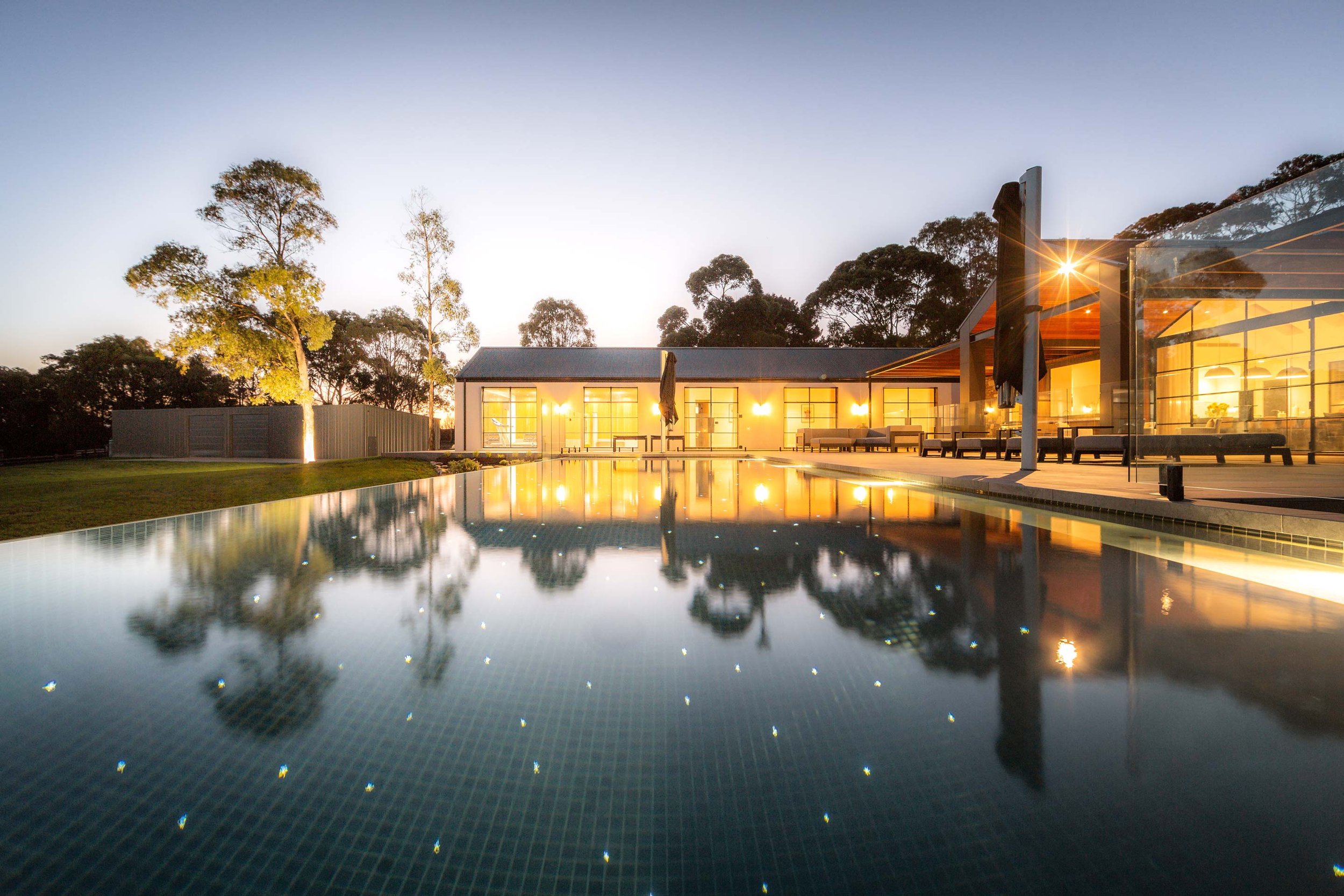 A wet-edge swimming pool illuminated with subtle, starlight-like lighting, surrounded by a warm and beautifully lit modern home.