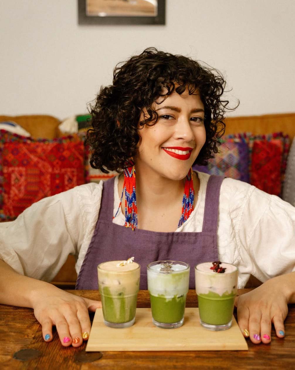 Smiling Latina entrepreneur and owner of Las Perras Cafe wearing a purple apron and colorful beaded necklace presenting specialty drinks at her Houston Latina-owned coffee shop