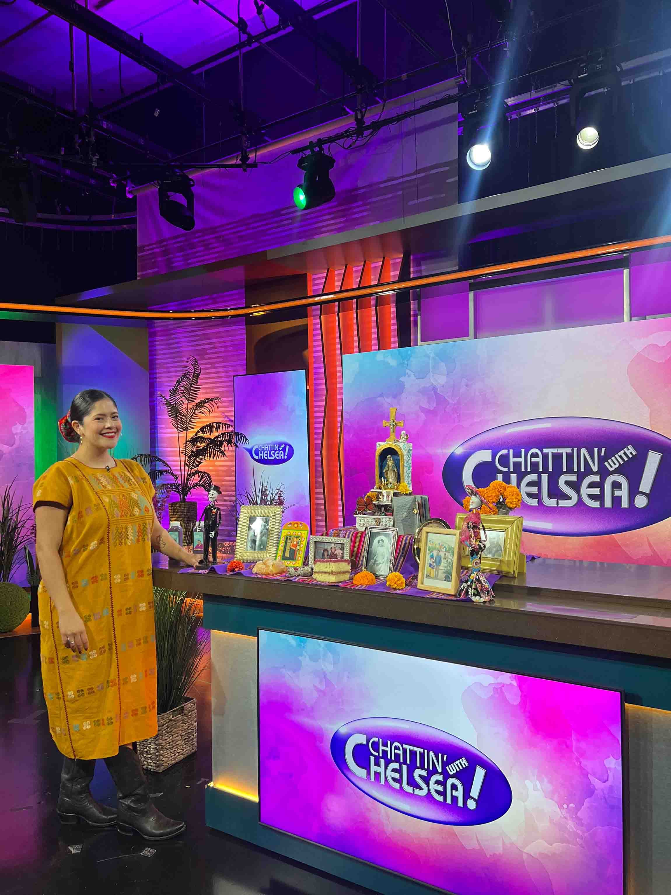 Latina entrepreneur in a traditional embroidered yellow dress presenting a Día de los Muertos ofrenda with marigolds and framed photos on the set of Chattin' with Chelsea, a Houston media appearance secured through multicultural PR strategy