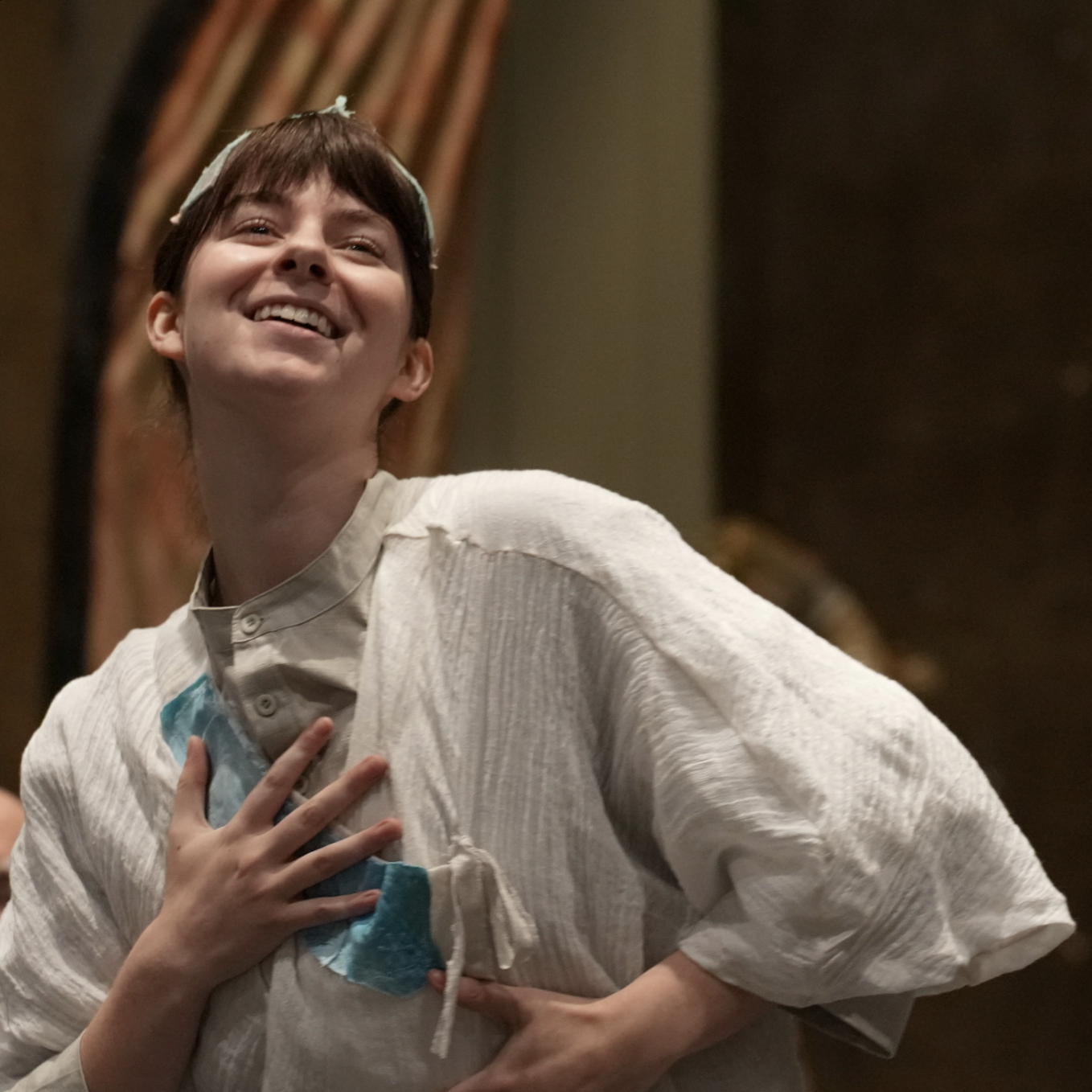 A person with short brown hair laughing, wearing a white garment with a stethoscope and a blue medical mask hanging from their neck, indoors with curtains in the background.