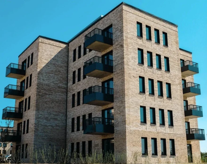 Lägenhetshus i ljus tegel med flera våningar, fotograferat utomhus under en klar ljusblå himmel.
