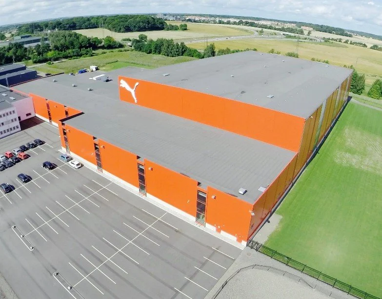 Flygvy över Puma-anläggning med stor industribyggnad, parkering och grönområden i Skåne.