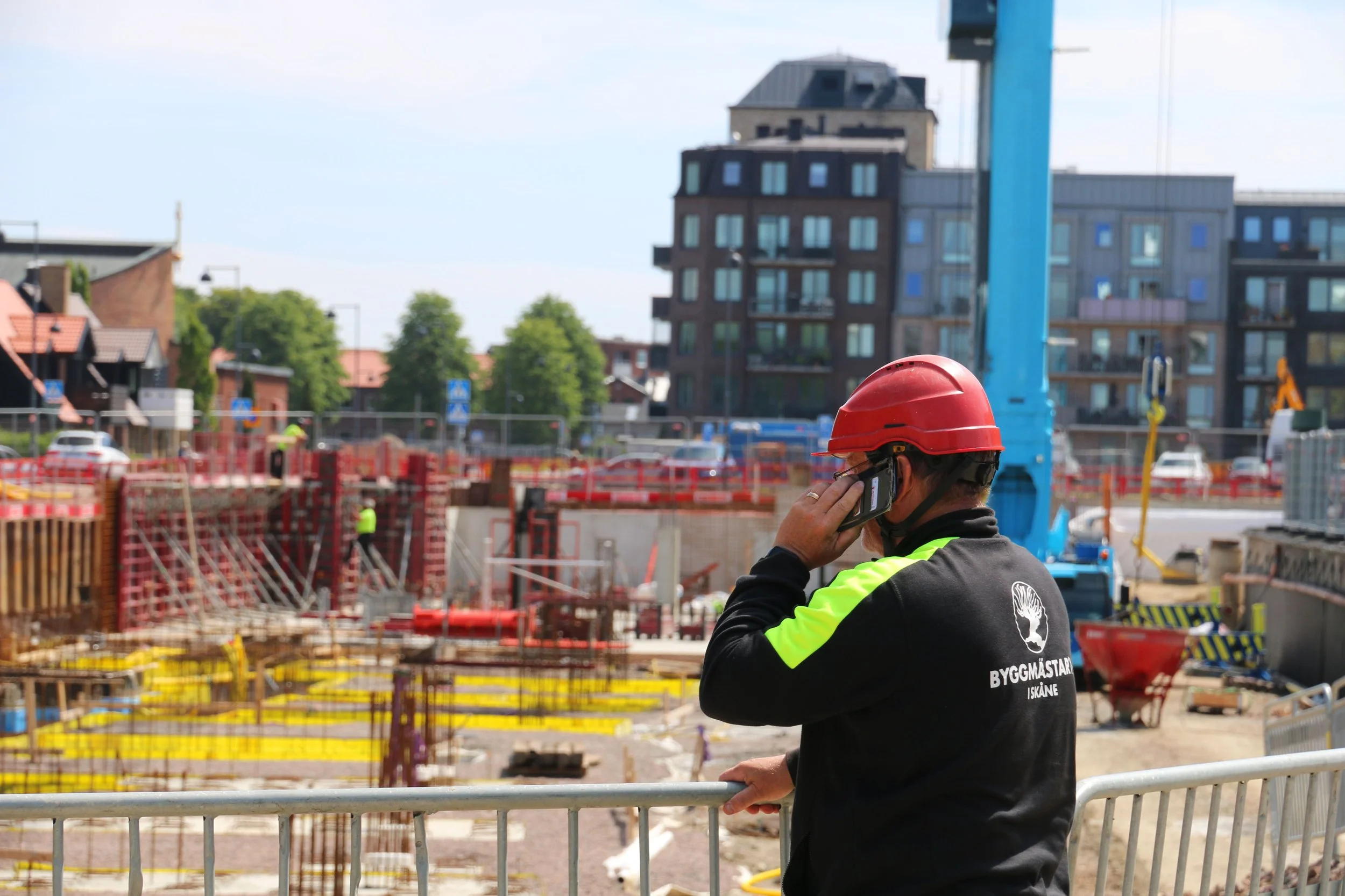Vraselklädd person md hjälm står ut över en byggarbetsplats och pratar i telefon.