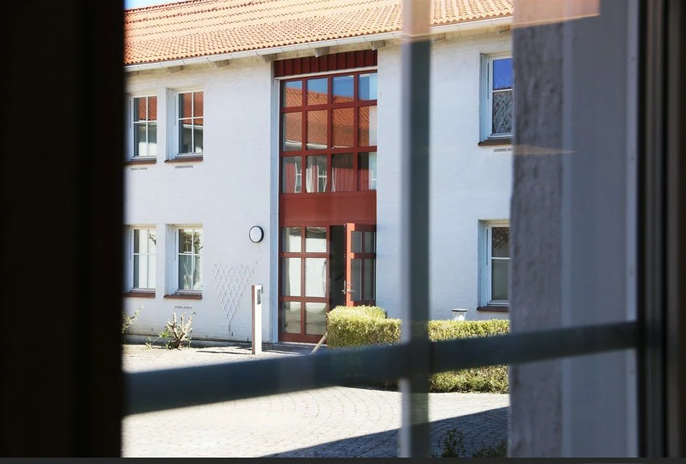 Vy genom ett fönster mot en byggnad med rödmålad entré, glaspartier och stenlagd gård.