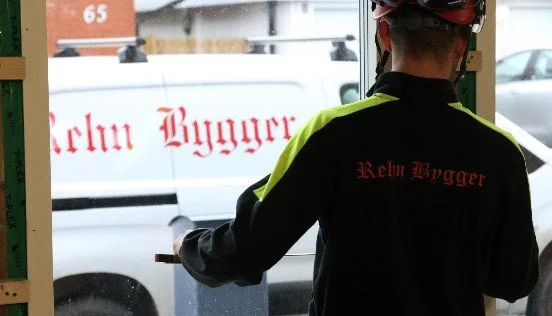 En hantverkare står med ryggen mot och mäter en fönsterkarm. Ute står en vit lätt skåpbil med en röd Rehn Bygger logotyp.