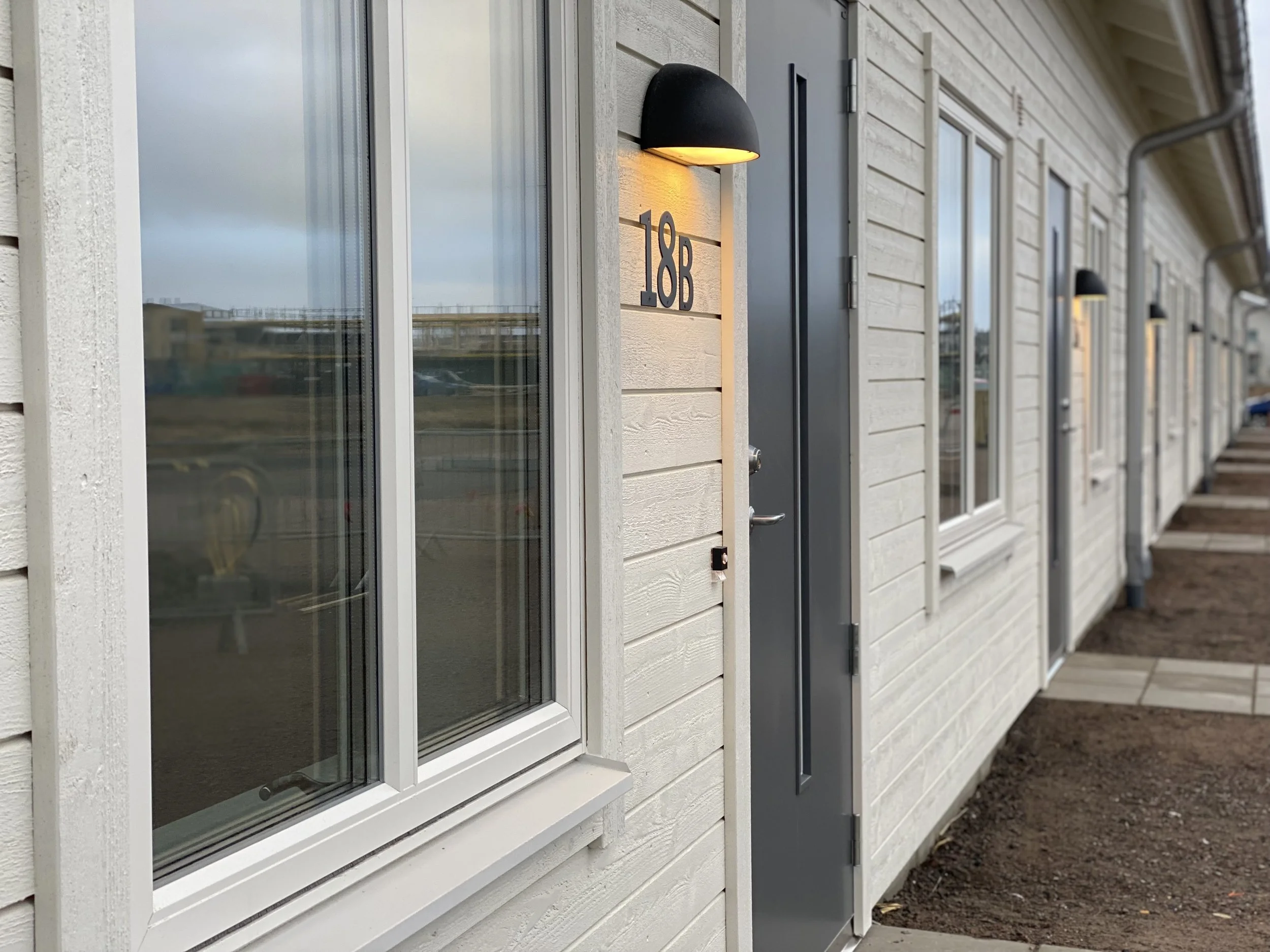 En vit trädfasad med en grå dörr, där en lampa lyser ovanför husnumret 18 B.