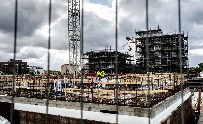 Byggarbetsplats med flera byggnader under konstruktion, byggnadsställningar och tre arbetare i reflekterande jackor, inhägnad med svart staket.