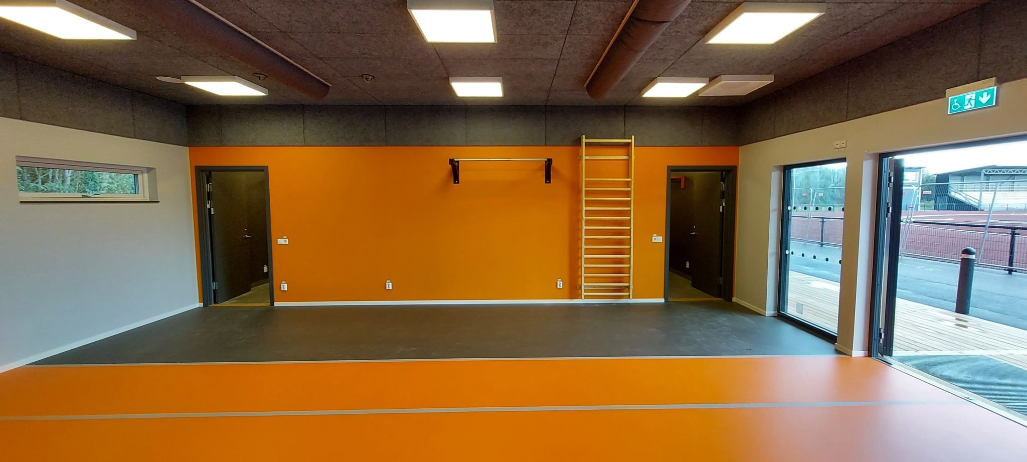 Ljust rum med stora fönsterpartier, oranget golv och orange vägg, en ribbstol i trä samt en väggmonterad stång.