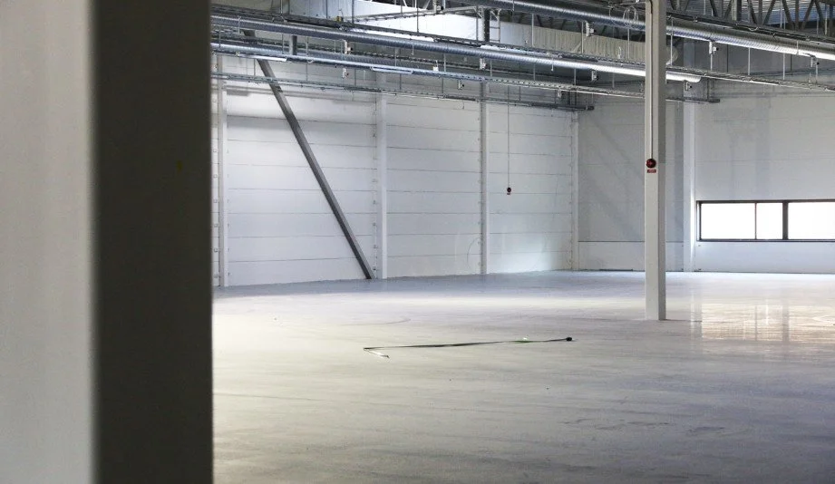 Tom industrilokal med högt i tak, betonggolv och synliga installationer i taket.