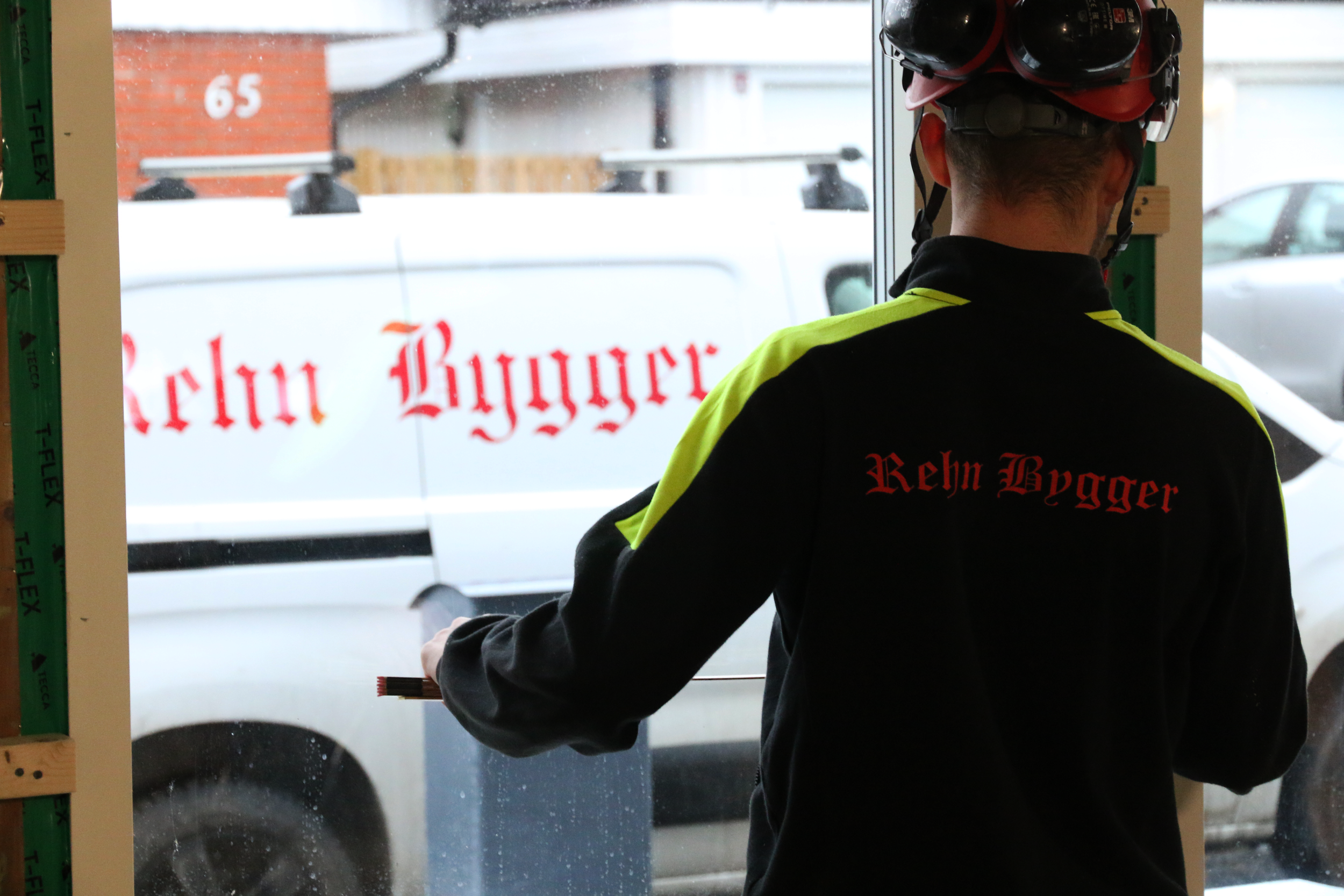 En hantverkare står med ryggen mot och mäter en fönsterkarm. Ute står en vit lätt skåpbil med en röd Rehn Bygger logotyp.