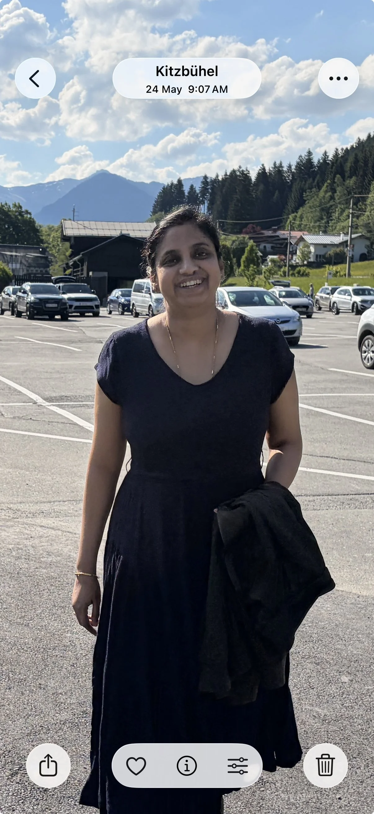 A woman smiling outdoors in a parking lot with mountains and trees in the background, bright sky with clouds, wearing a black dress, holding a jacket, and standing in direct sunlight.