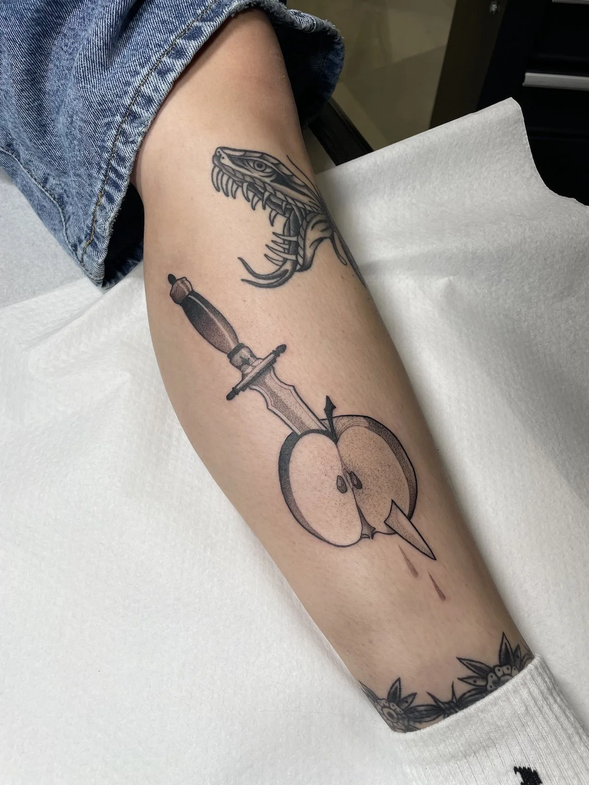 Tattoo of a dragon's head, a sword, and an apple with a lightning bolt piercing through it on a person's forearm.