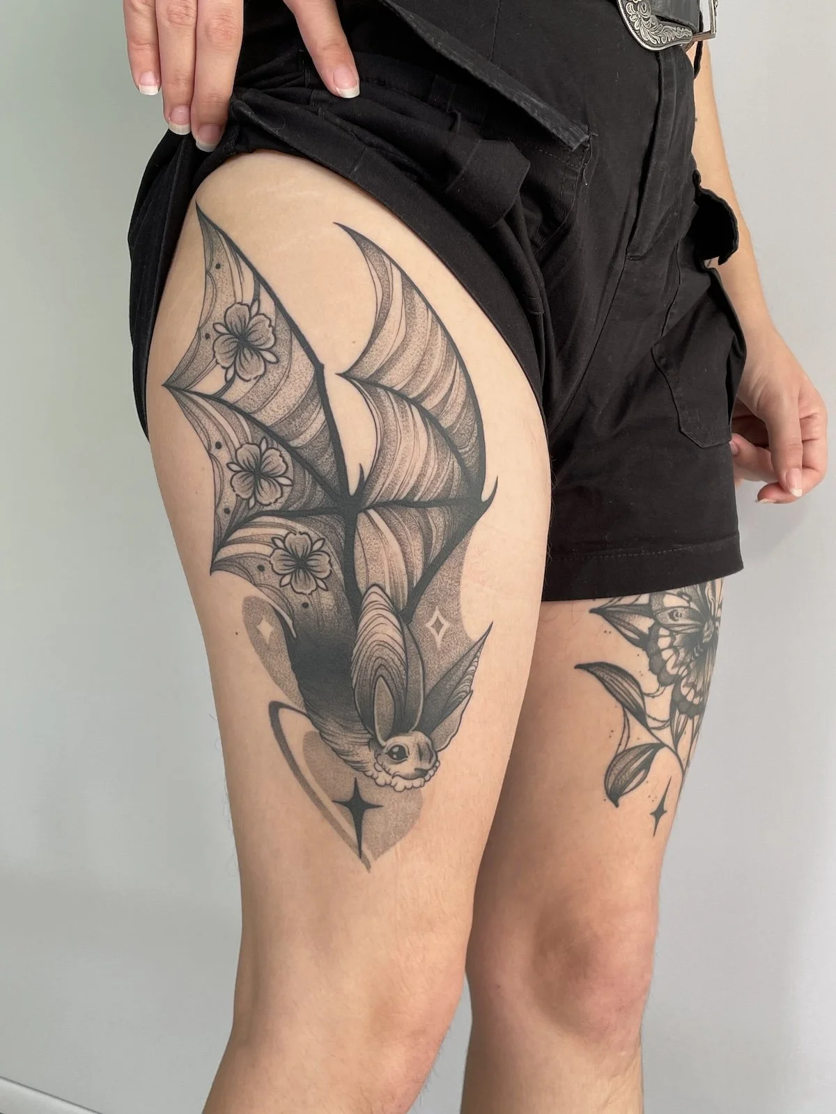 Tattoo of a bat with flowers and stars on the upper thigh of a person wearing black shorts.