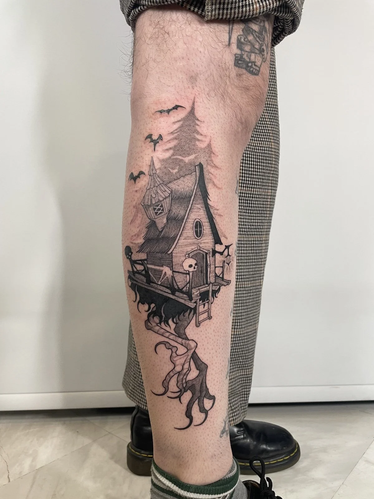 Tattoo of a haunted house with bats, a skull, and swirling smoke on a person's leg.