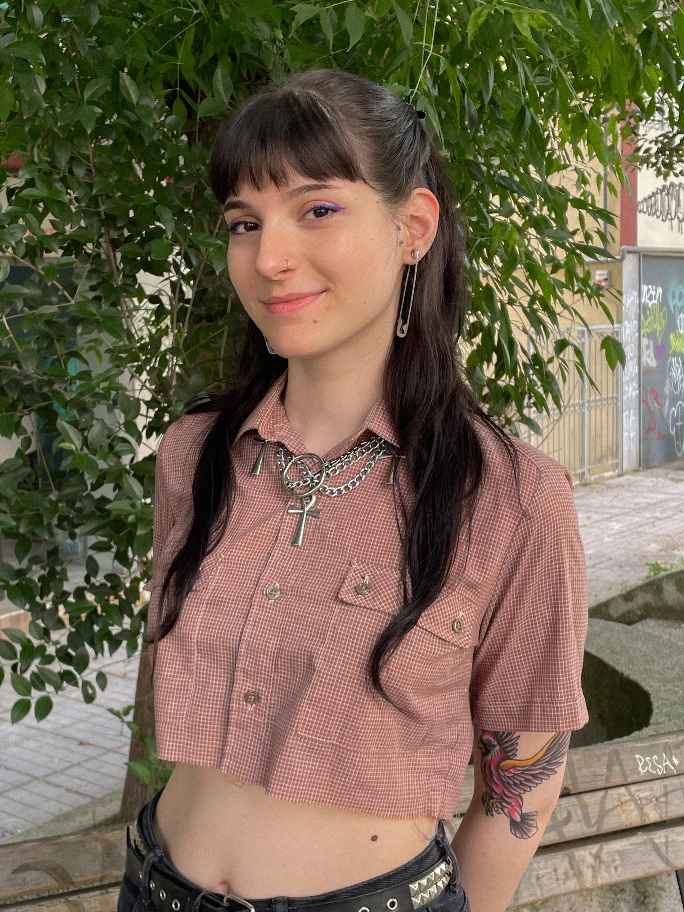 Young woman with dark hair and bangs, wearing purple eyeliner, a nose ring, layered chains with a cross pendant, a crop top, and a tattoo of a colorful bird on her arm. She is standing outdoors in front of green leaves with a graffiti-covered wall in the background.