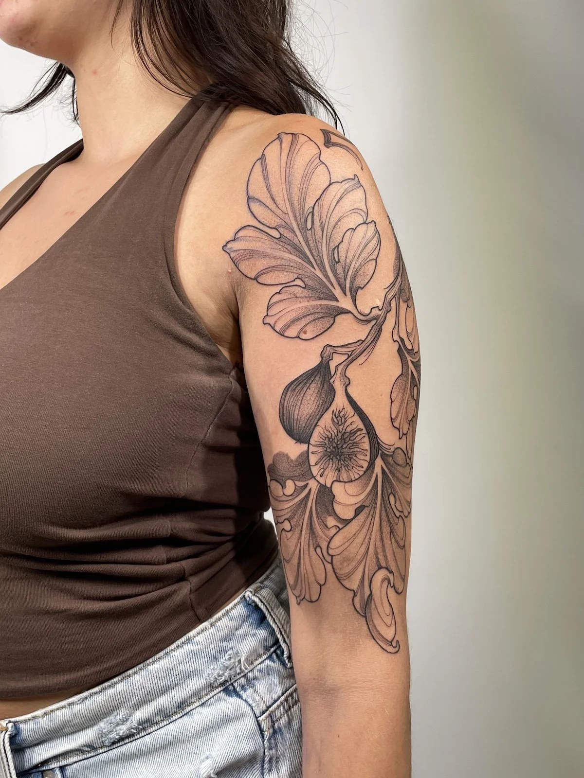 A woman with a detailed black line floral tattoo on her upper arm and shoulder, wearing a brown sleeveless top and light denim jeans.
