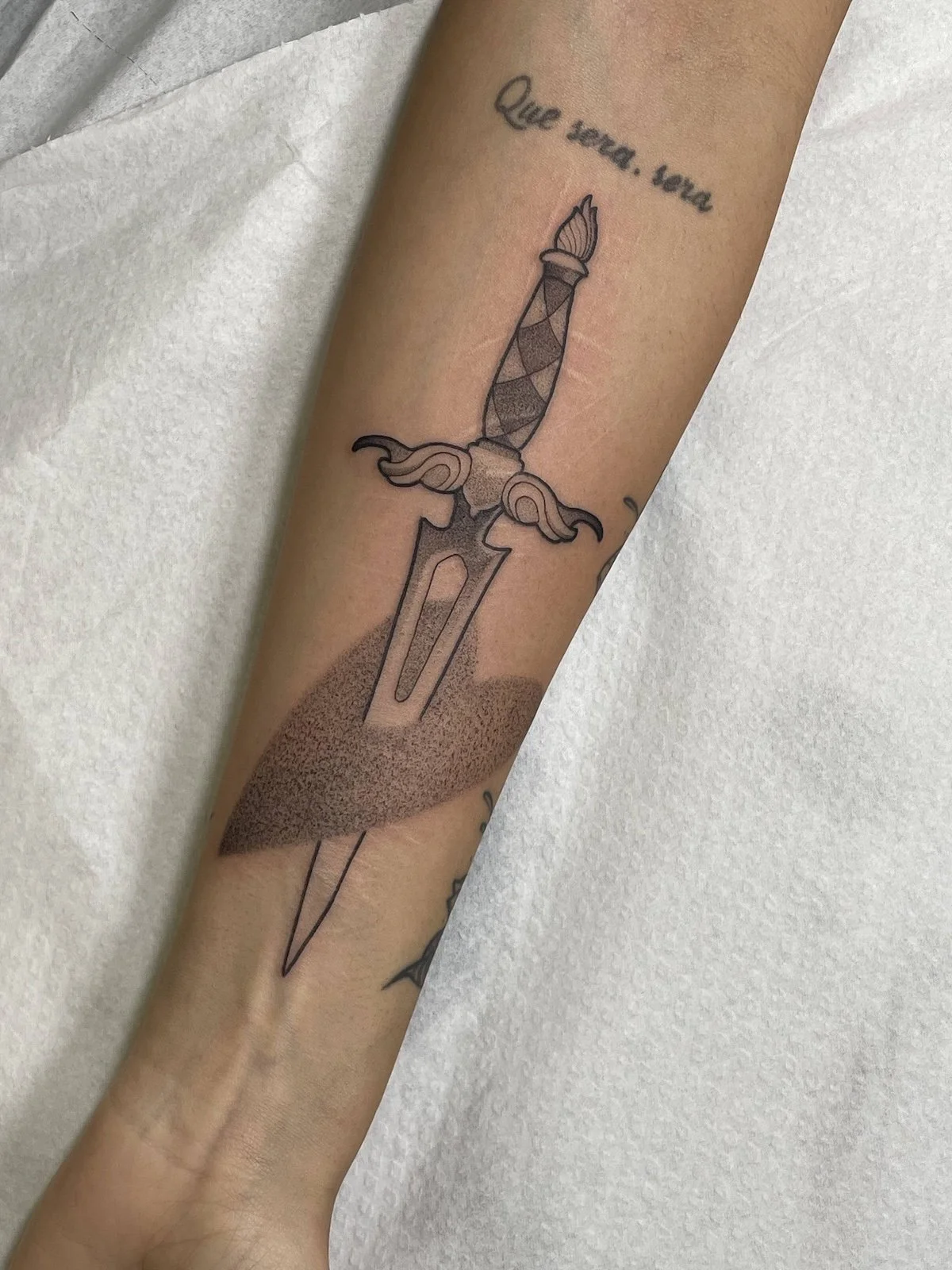 Tattoo of a dagger with a textured blade, a decorative hilt with wings, and a tattooed phrase in cursive, 'Que seas, sea', on a person's forearm.