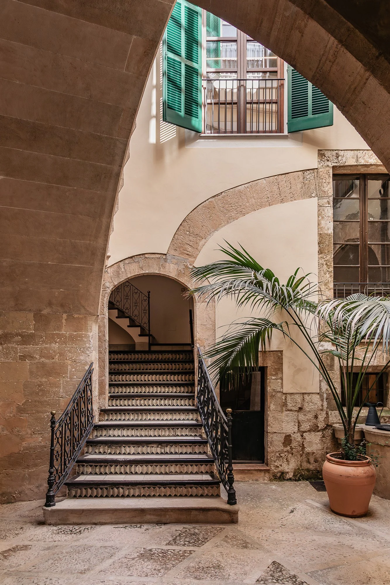 Interior shot of a historical building courtyard with stone stairs, green shutters on glass doors, a large potted palm, and an arched stone doorway.