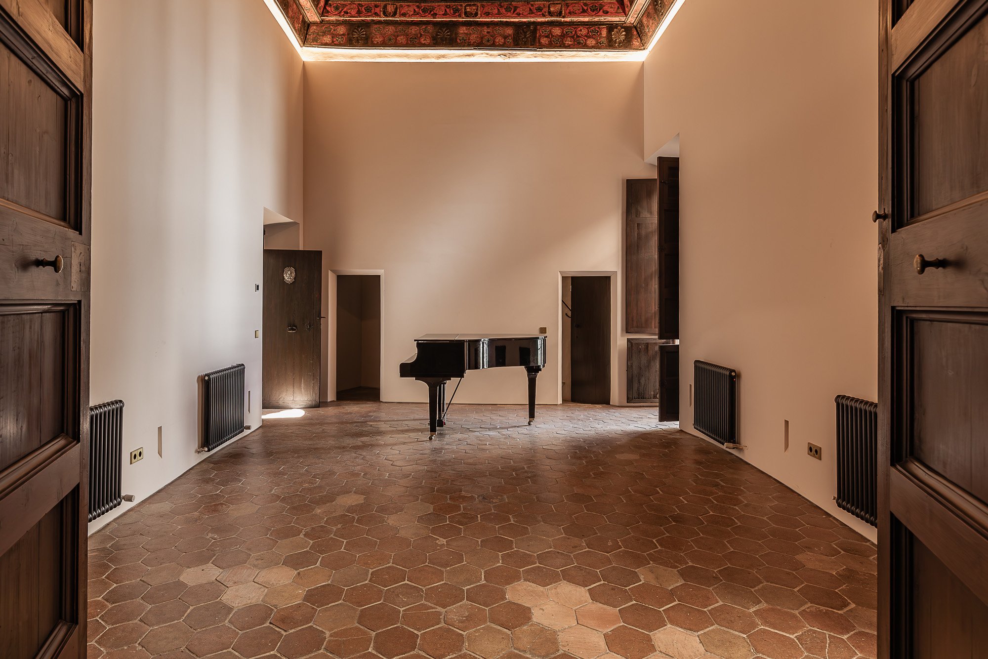 An empty music room with a grand black piano centered on a hexagonal terracotta tile floor, white walls, wood-paneled doors, built-in shelves on the right, hot water radiators, and a high ceiling with decorative painted beams.